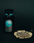 A dark glass jar with a blue and brown label labeled "Quintessence™ Tea for Opening & Healing the Throat Chakra" by Magic Hour sits next to a flower-shaped dish filled with a dried herbal mixture of organic loose leaf tea. A small spoon rests on the dish, all set against a dark background.