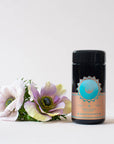 A black cylindrical container of Quintessence™ Tea for Opening & Healing the Throat Chakra with a pink and blue label sits next to a small bouquet of pale pink and purple flowers. The label shows the brand name "Magic Hour" and other text, with a turquoise emblem at the top. The background is plain white.