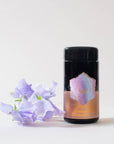 A black cylindrical tea canister with a pastel floral design and "Lotus Mind" text stands on a white surface. Beside it, there is a stem of light purple flowers. The background is plain white, highlighting the elegance of this Magic Hour Lotus Mind™ Tea of the Crown Chakra Traveler canister containing premium loose leaf tea.