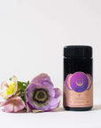 A black cylindrical container labeled "Blissful Black Teas Medley" stands on a light surface next to a cluster of pastel-colored flowers. The container, designed with light peach and purple accents, prominently features text and the Magic Hour logo. This elegant packaging hints at an exquisite blend of organic tea inside. The background is plain white.