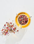 A cup of organic tea with loose leaf tea leaves and petals scattered around it on a white, textured surface. The tea is in a glass cup with a metal infuser, and the scattered leaves include various colorful dried herbs and flowers, reminiscent of Magic Hour Classics - Luxe Sampler Sips.