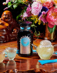 A dark bottle with an ornate label is placed on a wooden surface beside two glass cups, a clear teapot filled with light yellow organic tea, a strand of blue beads, and a colorful bouquet of flowers. A statue is visible in the background. The label reads "Quintessence™ Tea for Opening & Healing the Throat Chakra" by Magic Hour.