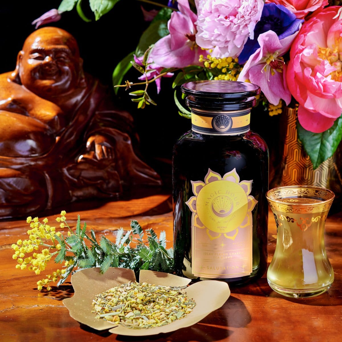 A serene setup featuring a jar of Salutation™ Tea for the Solar Plexus Chakra with a golden label, a small glass filled with brewed tea, and a paper holding loose organic ingredients. Surrounding this are vibrant flowers in a vase and a smiling Buddha statue, all placed on a wooden surface.