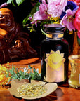 A serene setup featuring a jar of Salutation™ Tea for the Solar Plexus Chakra with a golden label, a small glass filled with brewed tea, and a paper holding loose organic ingredients. Surrounding this are vibrant flowers in a vase and a smiling Buddha statue, all placed on a wooden surface.