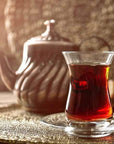 A glass of Bohemian Breakfast™ Violet Glass Traveler from Magic Hour sits on a clear saucer, with a traditional teapot in the background. The deep reddish-brown color of the organic tea is richly inviting, and the scene is warmly lit, creating a cozy atmosphere.