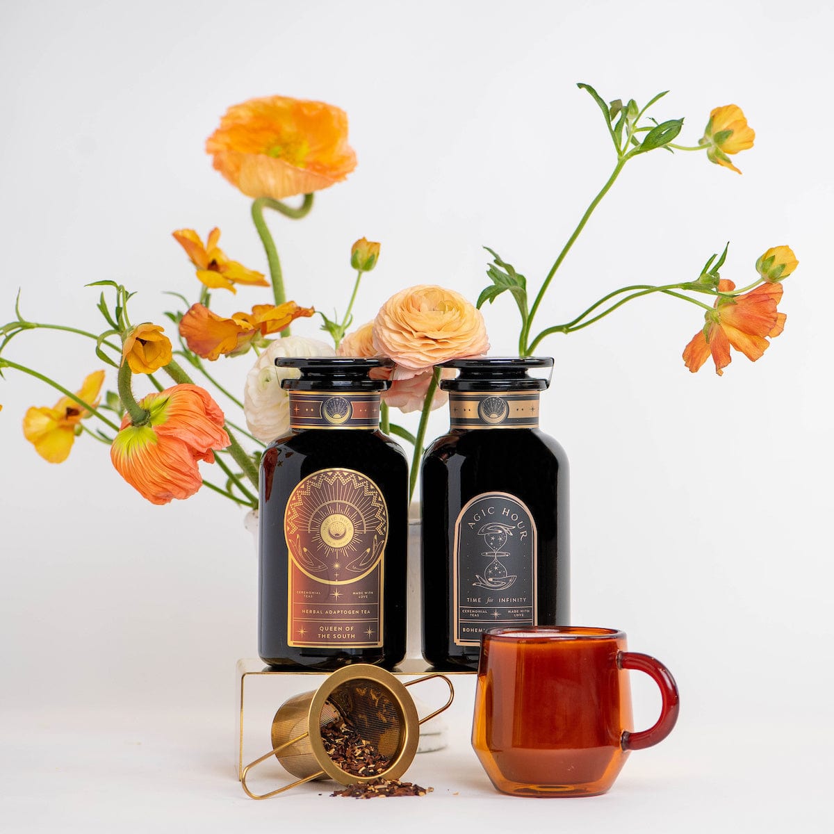 Two dark glass bottles with ornate labels stand on a small platform surrounded by bright orange and yellow flowers. A red glass cup and a gold measuring spoon containing organic tea ingredients sit in the foreground, creating an aesthetic still life arrangement that suggests these might be from the Coffee Lovers Tea Kit for Better Moods, Digestion & Metabolism.