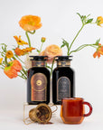 Two dark glass bottles with ornate labels stand on a small platform surrounded by bright orange and yellow flowers. A red glass cup and a gold measuring spoon containing organic tea ingredients sit in the foreground, creating an aesthetic still life arrangement that suggests these might be from the Coffee Lovers Tea Kit for Better Moods, Digestion & Metabolism.