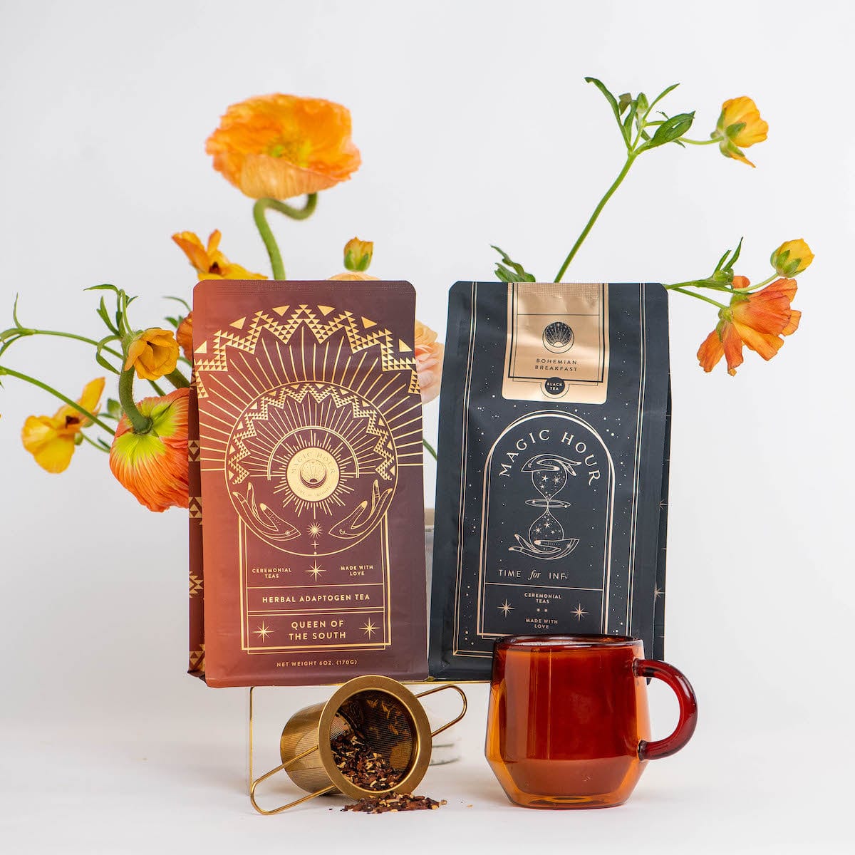 Two tea packages, one titled "Queen of the South" and the other "Magic Hour", are displayed against a white background with orange flowers. A transparent brown teacup and a gold tea infuser with loose organic tea ingredients complete the arrangement, showcasing the Coffee Lovers Tea Kit for Better Moods, Digestion & Metabolism as the perfect coffee alternative.