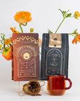 Two tea packages, one titled "Queen of the South" and the other "Magic Hour", are displayed against a white background with orange flowers. A transparent brown teacup and a gold tea infuser with loose organic tea ingredients complete the arrangement, showcasing the Coffee Lovers Tea Kit for Better Moods, Digestion & Metabolism as the perfect coffee alternative.