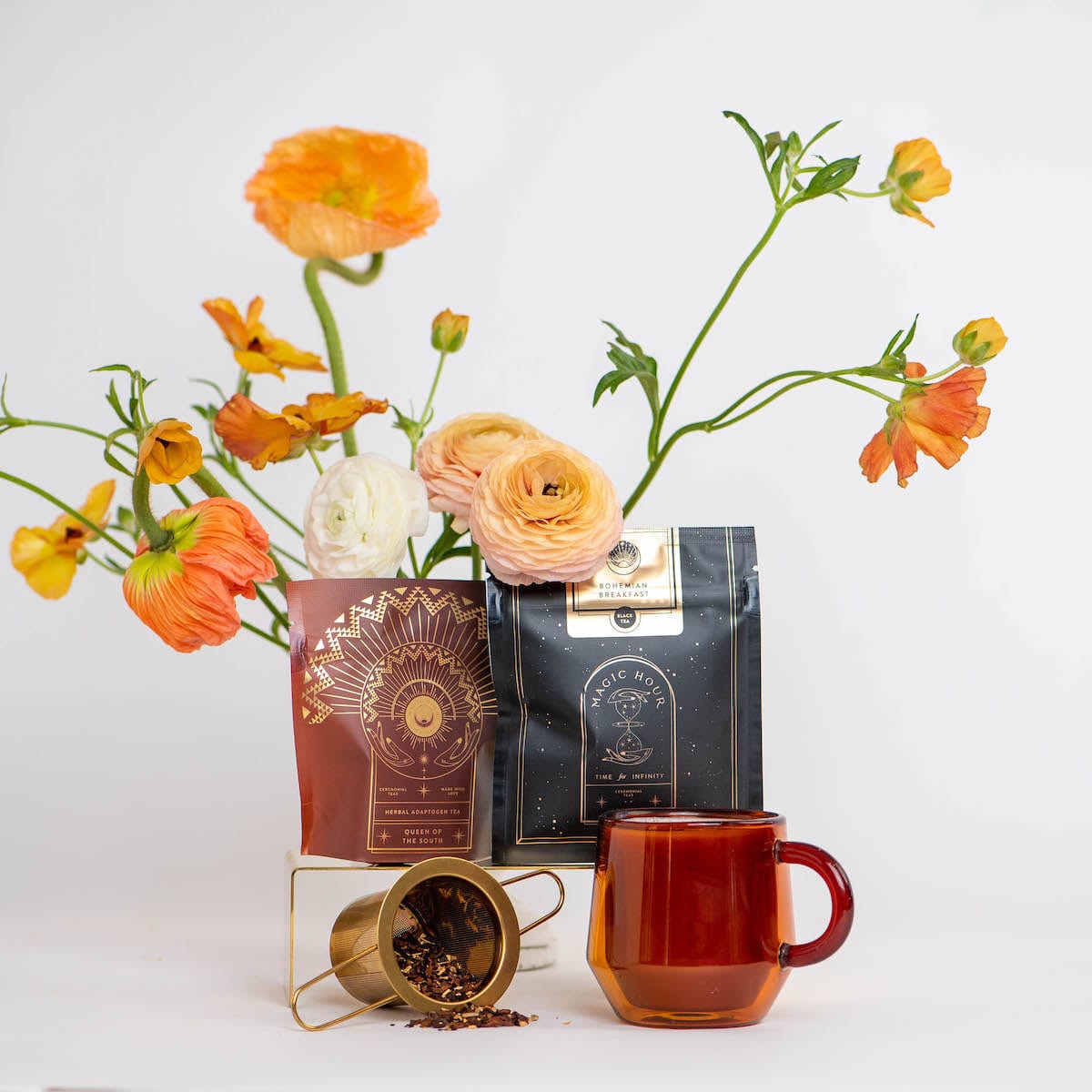 A setup with vibrant orange and white flowers in the background, two aesthetically designed packages from the Coffee Lovers Tea Kit for Better Moods, Digestion & Metabolism in the foreground—one black and gold, the other red and gold—showcasing mood-boosting teas, a small metal container spilling organic tea ingredients, and a red glass mug.