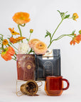 A setup with vibrant orange and white flowers in the background, two aesthetically designed packages from the Coffee Lovers Tea Kit for Better Moods, Digestion & Metabolism in the foreground—one black and gold, the other red and gold—showcasing mood-boosting teas, a small metal container spilling organic tea ingredients, and a red glass mug.