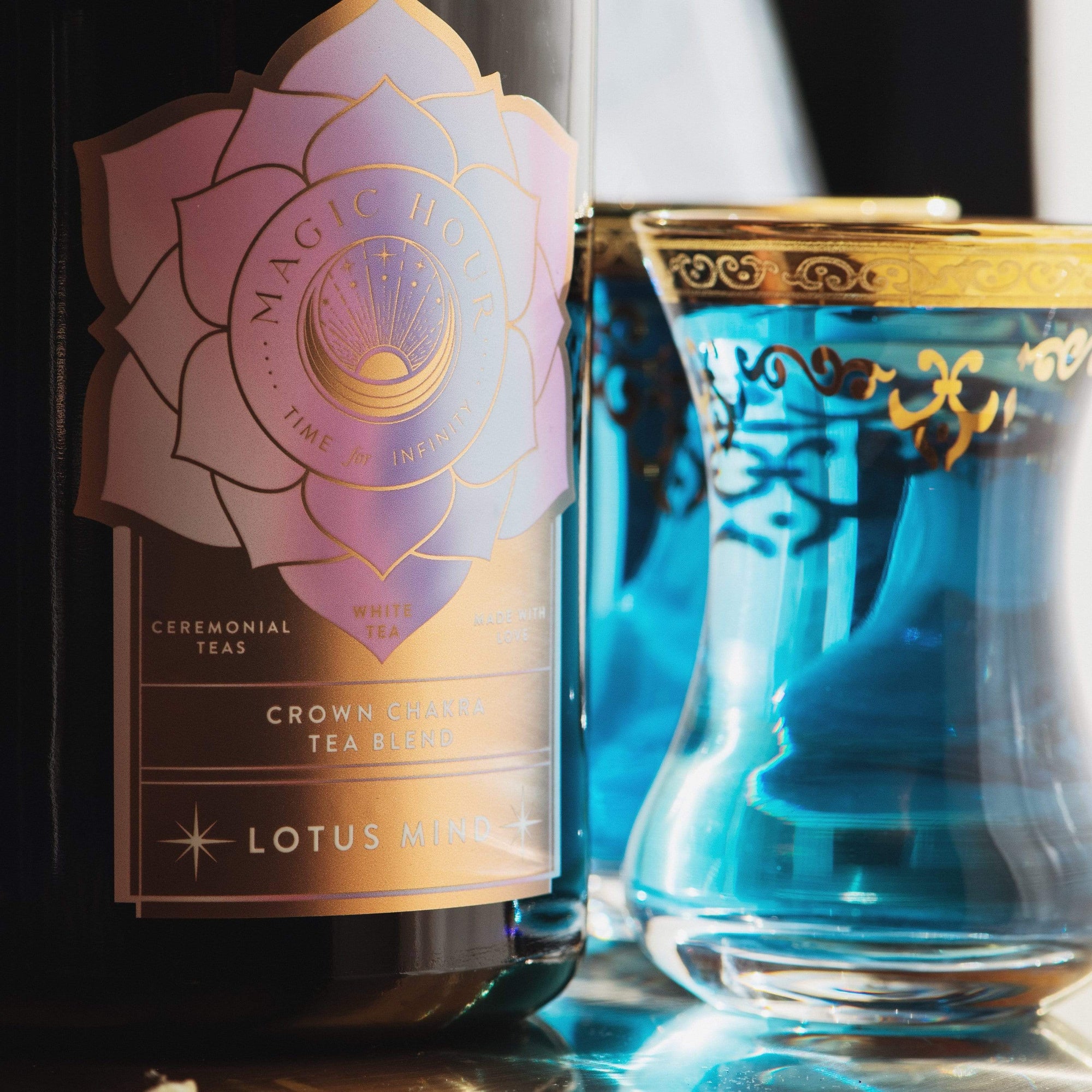 A close-up shot of a bottle labeled "Magic Hour" next to a glass filled with blue liquid. The label on the bottle reads "Lotus Mind™ Tea of the Crown Chakra Traveler." The glass, adorned with gold decorative accents along the rim and design, complements the allure of this organic, loose leaf tea.