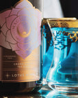 A close-up shot of a bottle labeled "Magic Hour" next to a glass filled with blue liquid. The label on the bottle reads "Lotus Mind™ Tea of the Crown Chakra Traveler." The glass, adorned with gold decorative accents along the rim and design, complements the allure of this organic, loose leaf tea.
