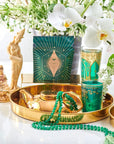 A decorative setup featuring a gold tray with a teapot, green beaded necklace, and ornate green cups. In the background are white flowers, greenery, a bag of Club Magic Hour Organic Ceremonial Matcha 700, and two statuettes of women. The organic tea enhances the fresh ambiance.
