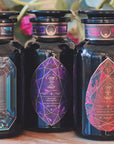 Three dark glass bottles with ornate, geometric labels are displayed side by side on a wooden surface. The labels feature intricate crystal designs in blue, purple, and red tones. A blurred background includes green leaves and a pink flower, enhancing the allure of the **Magic Hour Gemstone Bundle: Garnet, Amethyst, Aquamarine**.