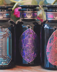 Three dark glass bottles with ornate, geometric labels are displayed side by side on a wooden surface. The labels feature intricate crystal designs in blue, purple, and red tones. A blurred background includes green leaves and a pink flower, enhancing the allure of the **Magic Hour Gemstone Bundle: Garnet, Amethyst, Aquamarine**.