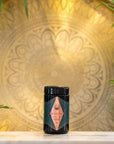 A black container with an ornate label is placed on a white surface. The background features a large, intricate, circular golden mandala design with ambient lighting. Green leaves partially frame the image on the left and right sides, highlighting the allure of Magic Hour Tea Connoisseur Collection inside.