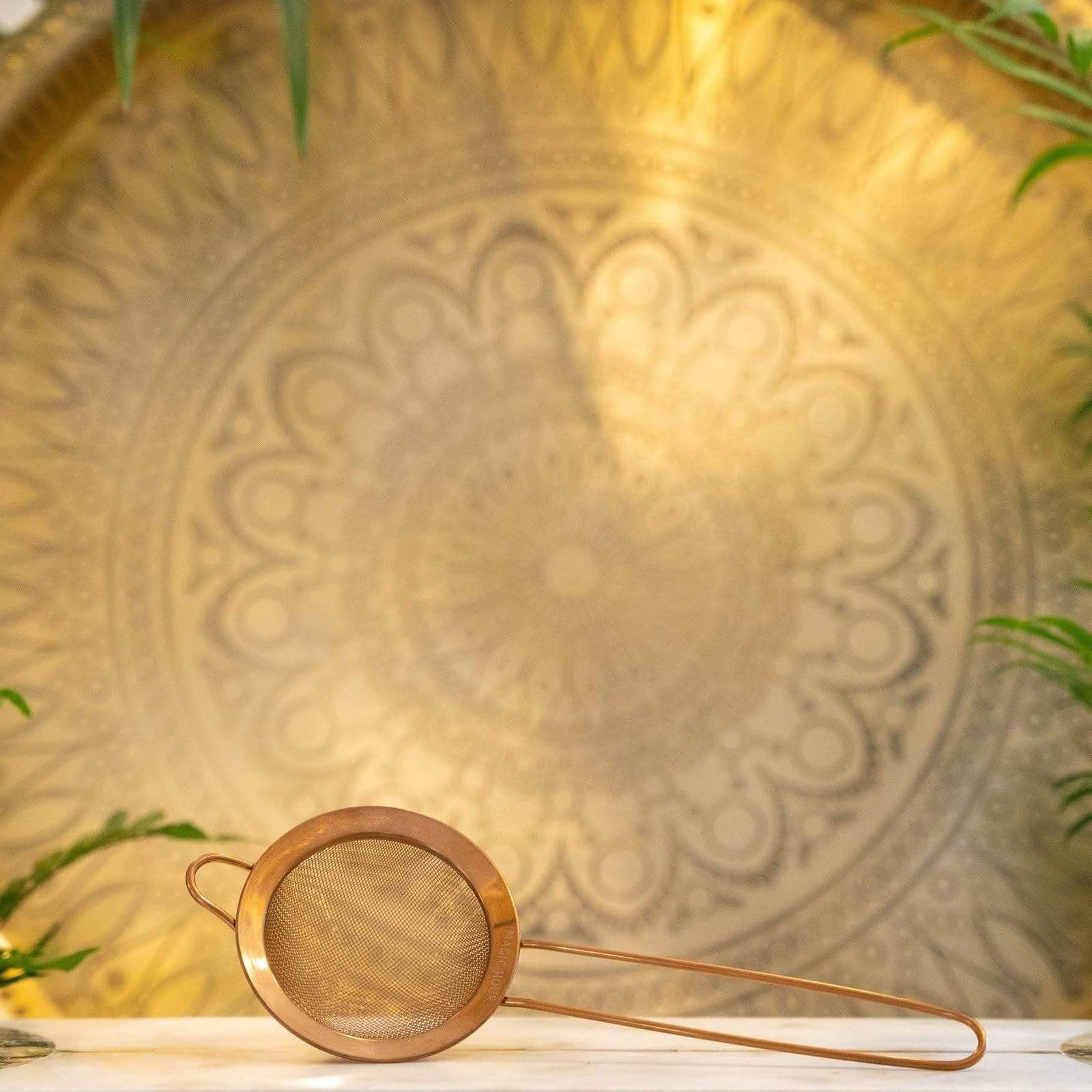 A Ceremonial Matcha Traveler Gift Box from Magic Hour is positioned on a white surface in the foreground. The background features an intricate, circular mandala pattern illuminated by warm, ambient lighting. Green plant leaves frame the edges of the image, evoking the serene ritual of Loose Leaf Tea preparation.