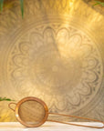 A Ceremonial Matcha Traveler Gift Box from Magic Hour is positioned on a white surface in the foreground. The background features an intricate, circular mandala pattern illuminated by warm, ambient lighting. Green plant leaves frame the edges of the image, evoking the serene ritual of Loose Leaf Tea preparation.