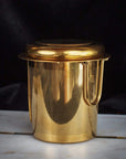 A gold-toned metallic tea infuser with a cylindrical shape and a perforated body, featuring a fitted lid and a handle, is perfect for brewing your favorite Loose Leaf Tea. The PMB Mesh Screen Company's Midas Touch: Golden-Hued Tea Strainer is placed against a dark background on a light-colored surface.
