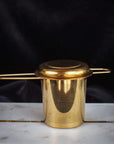 A Midas Touch: Golden-Hued Tea Strainer by PMB Mesh Screen Company, designed for loose leaf tea with a lid and two side handles, is placed on a white marble surface against a dark background. The tea strainer has a cylindrical shape with fine mesh sides, perfect for brewing your favorite organic tea.