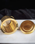 A Midas Touch: Golden-Hued Tea Strainer by PMB Mesh Screen Company, perfect for brewing Organic Tea, is displayed on a marble surface against a black background. The strainer’s fine mesh is ideal for Loose Leaf Tea. Nearby, its matching gold lid rests elegantly on the marble.
