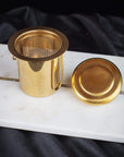 A gold-colored Midas Touch: Golden-Hued Tea Strainer by PMB Mesh Screen Company with fine mesh and a matching lid is displayed on a white marble surface with a dark fabric backdrop. Perfect for steeping your favorite loose leaf tea, the strainer stands upright, with the lid placed beside it.