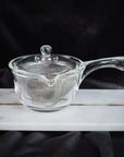 A small transparent glass teapot, the Tea-in-Hand: The Perfect Steep Side-pour Ceremonial Teapot by Espresso Parts, with a lid and a metal strainer inside, sits on a rectangular white marble slab. The background is dark and fabric-like, emphasizing the teapot's clear material and simple design, perfect for steeping your favorite loose leaf tea.