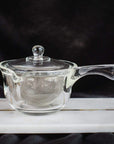 A small clear glass teapot with a lid and spout, featuring a metal mesh infuser inside, is placed on a white surface against a dark background. Perfect for brewing Magic Hour Tea, the Tea-in-Hand: The Perfect Steep Side-pour Ceremonial Teapot by Espresso Parts has a handle on one side and appears elegant and minimalist.