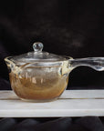 A transparent glass teapot with a long handle and a round lid is placed on a white rectangular marble base against a dark background. The Espresso Parts Tea-in-Hand: The Perfect Steep Side-pour Ceremonial Teapot, containing Organic Tea, reveals a light brown liquid with a mesh strainer visible inside.