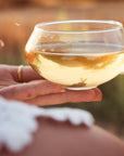 A close-up of a person holding a small, clear glass cup filled with light-colored Ti Quan Yin Oolong: Tea of Patient Compassion by Magic Hour. Another person's hand is gently holding the cup's handle. The person in the foreground is wearing a white, lace outfit, and the background is blurred with warm, natural colors.