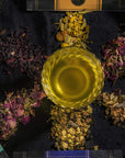A glass of yellow-colored tea is placed at the center of the image, surrounded by six piles of loose leaf teas and herbs in different colors and textures. The Self-Guided 8 Day Ceremonial Tea Cleanse Kit lies nearby, ready to guide you on an 8-day