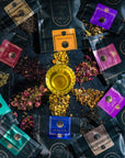 A glass cup filled with yellow liquid stands at the center of a dark background, surrounded by an assortment of colorful tea packets from the Self-Guided 8 Day Ceremonial Tea Cleanse Kit. Loose tea leaves and petals are scattered around, blending harmoniously with the 8-day