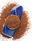 A circular pile of finely ground Rise & Shine Herbal Tea leaves from Club Magic Hour is placed on a blue stone. The blue stone serves as a base, with the tea leaves evenly spread over it and scattered around, creating an earthy and textured visual against a white background.