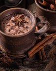 A rustic wooden mug filled with Queen of the South: Delicious Cocoa Detox Tea, garnished with star anise and chocolate shavings. Next to the mug are cinnamon sticks and another mug filled with whole cinnamon sticks, set on an aged wooden surface, creating a cozy ambiance worthy of a Magic Hour experience.