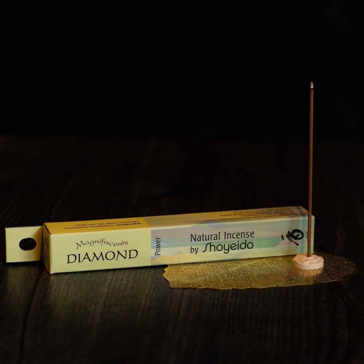 A horizontal box of Diamond Incense from Shoyeido rests on a dark wooden surface. To the right of the box, an incense stick rich with sandalwood and other fragrances is lit and held upright in a small holder, with a delicate golden mesh beneath it.