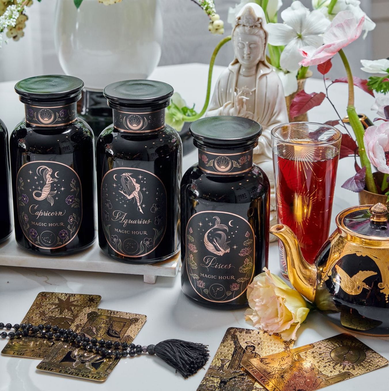A display featuring three black jars with astrological signs, surrounded by tarot cards, a brass teapot, beads, flowers, and a statue of a serene figure. The jars—labeled Capricorn, Aquarius, and Pisces—stand beside a glass of Magic Hour Tea's Astrology Bundle: Capricorn, Aquarius, Pisces creating an enchanting blend of elegance and mysticism.