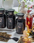 A display featuring three black jars with astrological signs, surrounded by tarot cards, a brass teapot, beads, flowers, and a statue of a serene figure. The jars—labeled Capricorn, Aquarius, and Pisces—stand beside a glass of Magic Hour Tea's Astrology Bundle: Capricorn, Aquarius, Pisces creating an enchanting blend of elegance and mysticism.