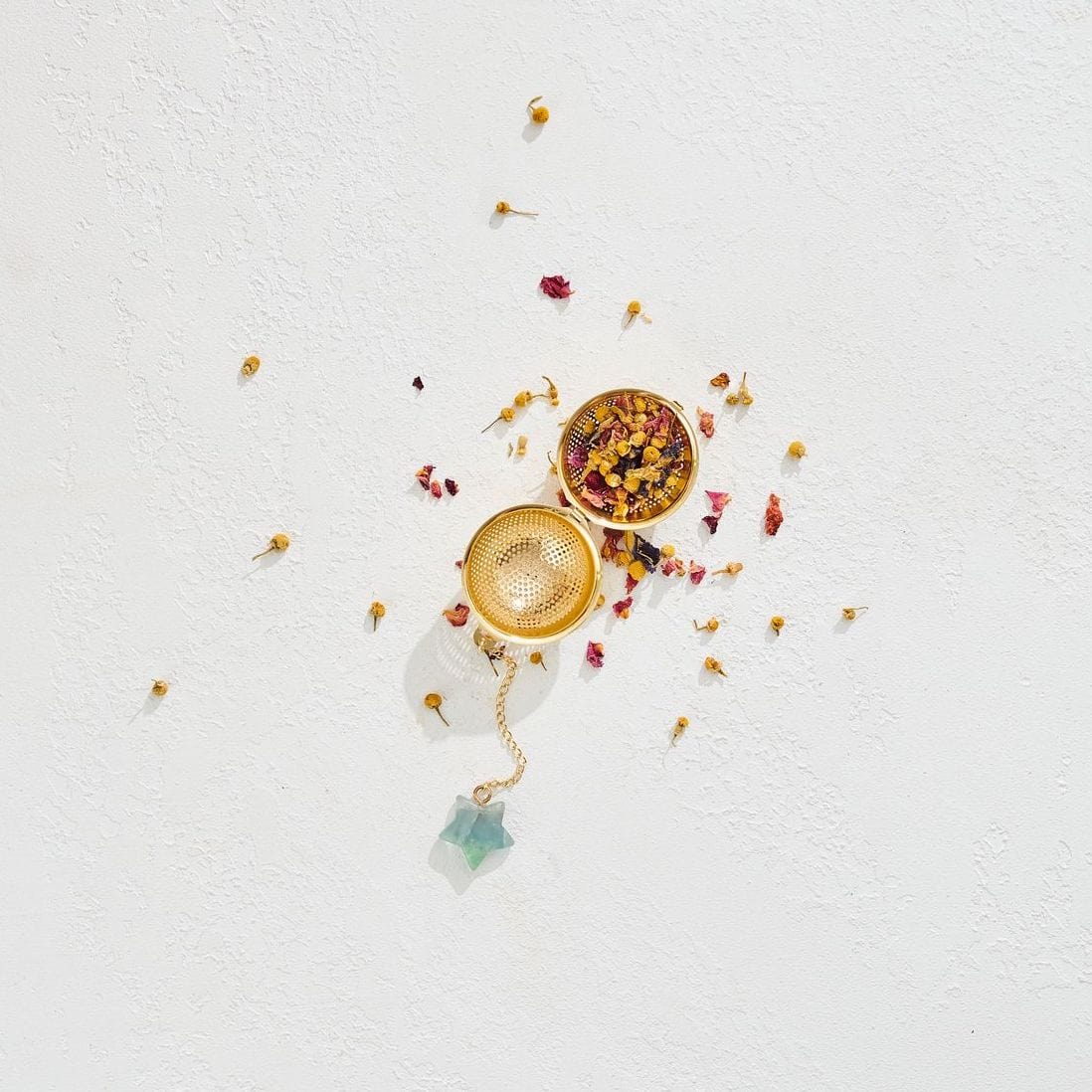 A gold Emerald Star Tea Strainer by Magic Hour with dried herbs and flowers scattered around it. The infuser is open, revealing more of the dried ingredients inside, and a small crystal is attached to the chain. The white, textured background highlights the colors of the organic loose leaf tea and gold strainer perfectly.