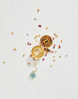 A gold Emerald Star Tea Strainer by Magic Hour with dried herbs and flowers scattered around it. The infuser is open, revealing more of the dried ingredients inside, and a small crystal is attached to the chain. The white, textured background highlights the colors of the organic loose leaf tea and gold strainer perfectly.