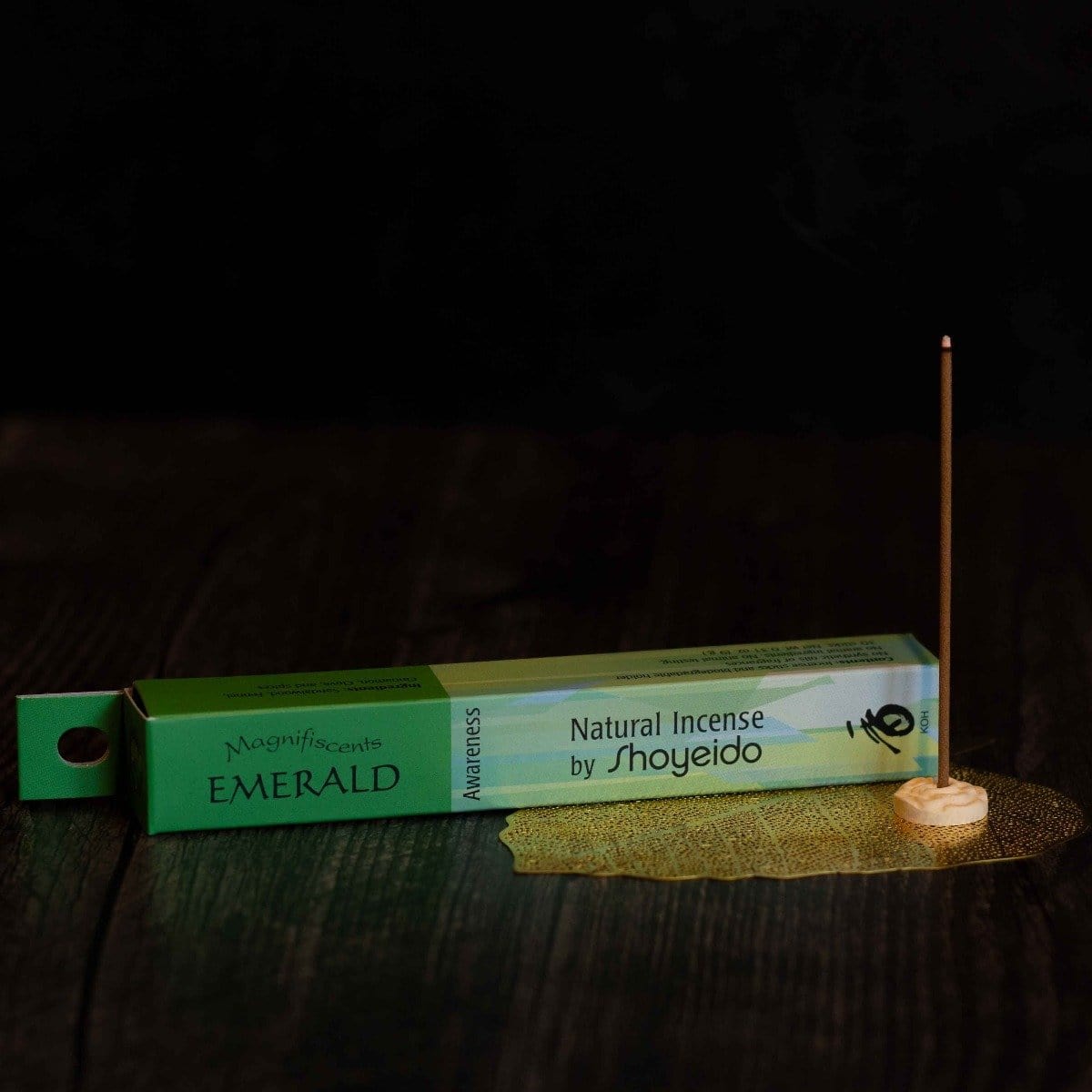 A Magic Hour natural incense stick named "Renew & Relax: Ritual Gift for Mindfulness & Calm" is lit and standing on a small holder. The incense packaging, green like Magic Hour Tea, rests on a dark surface. A delicate golden mesh is placed underneath the incense holder, creating an aura of tranquility and elegance.