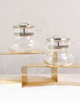 Two Essential Perfection Teapots with Built-in Strainers and metal lids are displayed on two yellow rectangular stands of different heights. These teapots, featuring built-in strainers, have a simple, modern design with round bodies, curved handles, and small spouts—perfect for enjoying loose leaf tea against the plain white fabric background.