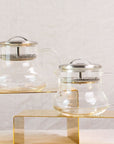 Two Essential Perfection Teapots with Built-in Strainers and metal lids are displayed on two yellow rectangular stands of different heights. These teapots, featuring built-in strainers, have a simple, modern design with round bodies, curved handles, and small spouts—perfect for enjoying loose leaf tea against the plain white fabric background.