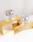 Two Essential Perfection Teapots, each featuring a built-in strainer and stainless steel lid, are displayed on gold-colored rectangular stands. Crafted from Borosilicate glass, they sit against a soft white fabric backdrop. Their simple, modern design exudes a minimalist and elegant aesthetic ideal for brewing loose leaf tea.