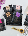 A collection of Magic Hour Classics - Luxe Sampler Sips packs arranged in a black box, with various flavors distinguished by pink, gold, and purple labels. A cup of organic tea sits nearby, along with a patterned golden tea scoop filled with loose leaf tea. Bright flowers are in the background.