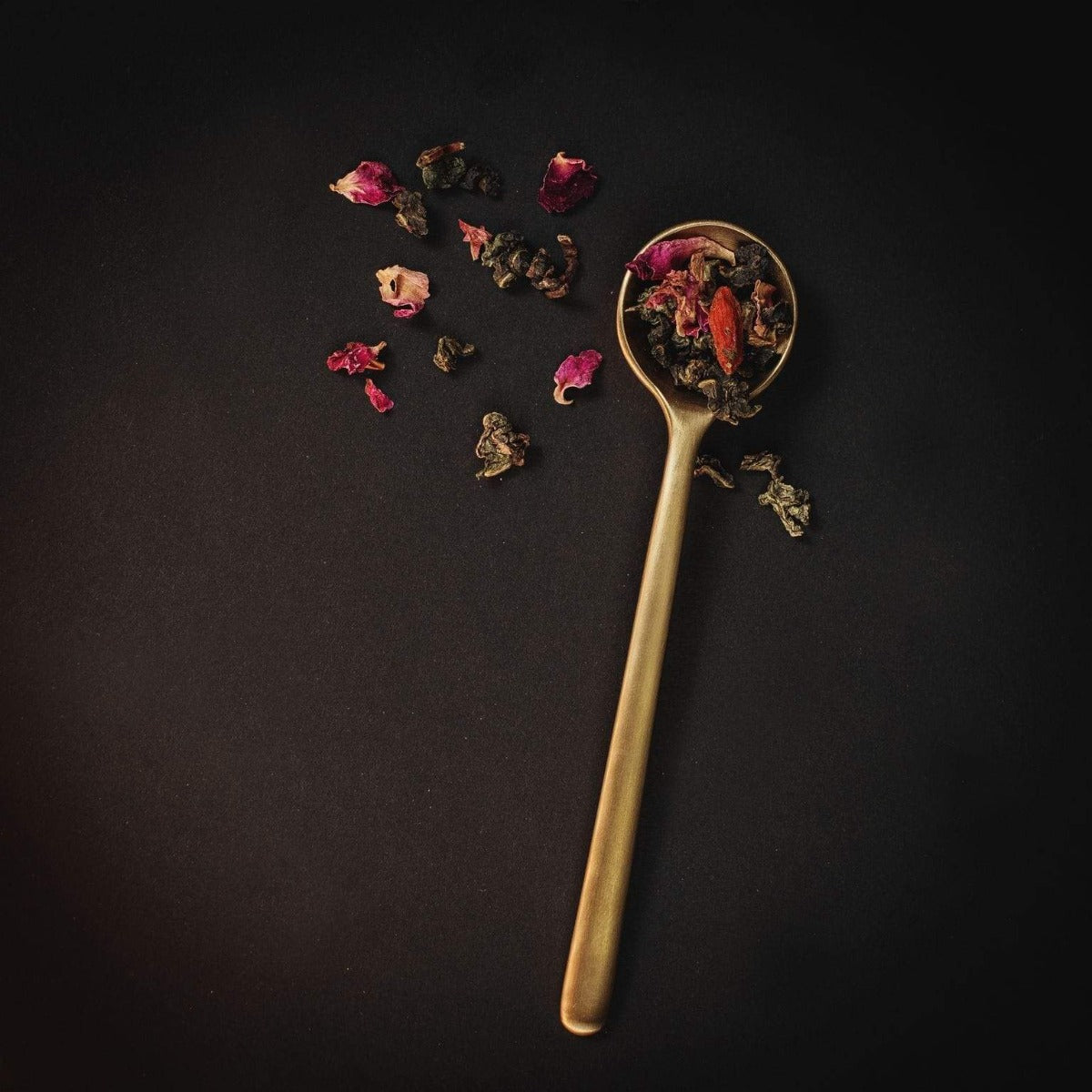 A golden spoon lies on a dark surface, filled with a variety of Renewal: Peach-Goji-Rose Oolong Tea leaves and flower petals by Magic Hour. Some of the loose leaf tea and petals are scattered around the spoon, creating an artistic arrangement against the black background.