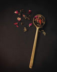 A golden spoon lies on a dark surface, filled with a variety of Renewal: Peach-Goji-Rose Oolong Tea leaves and flower petals by Magic Hour. Some of the loose leaf tea and petals are scattered around the spoon, creating an artistic arrangement against the black background.