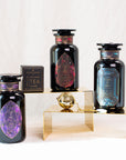 The image shows three black glass jars of Magic Hour Gemstone Bundle: Garnet, Amethyst, Aquamarine with colorful geometric labels, arranged on golden risers. Each label features intricate designs in different colors: purple, red, and blue. A small black box labeled "Magic Hour Tea Infuser" and a gold tea infuser are also displayed.