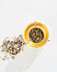 A clear glass cup filled with light yellow tea brewed from "Classics - Luxe Sampler Sips" sits on a white surface. A tea infuser with loose tea leaves rests atop the cup. To the left, a small pile of dry Coconut Chai leaves is scattered, ready to evoke memories of a serene Bohemian Breakfast.