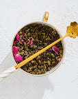 A cup filled with loose-leaf tea, adorned with pink flower petals, features the elegant Gold Floral Teaspoon resting across the top. The teaspoon, with its long gold stem and delicate white handle ending in a flower shape, is placed on a white textured surface.